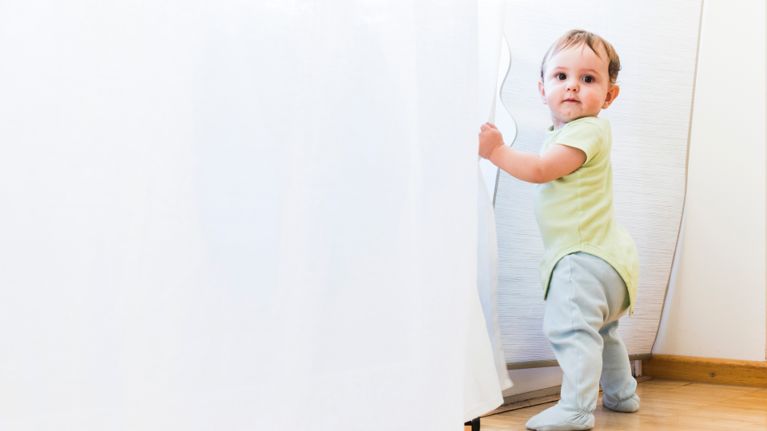 A baby in a lime green onesie standing and holding white curtains while looking at the camera