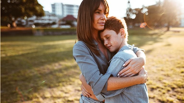 A mother stands in a sunlit park, holding a tween child close in a quiet, comforting hug.