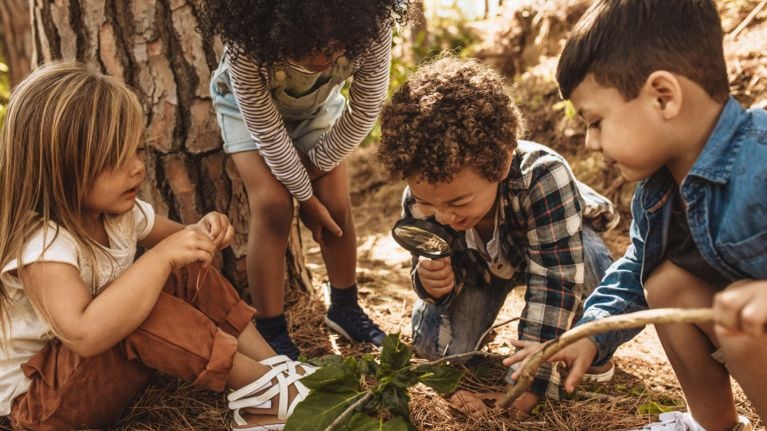 Four children crouch on a forest floor around a small plant, with one child peering through a magnifying glass as they explore together.
