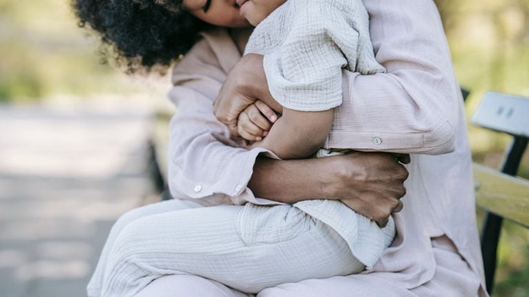 A parent sits outdoors on a bench, holding a young child close in their lap in a quiet, comforting embrace.