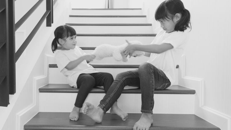 Two young children sit on a staircase, each pulling on the same stuffed toy while looking at each other, illustrating a moment of conflict over sharing.