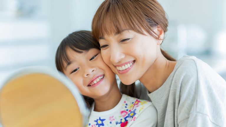 Smiling parent and child lean together and look into a mirror in a bright bathroom, suggesting a warm, everyday getting-ready moment.