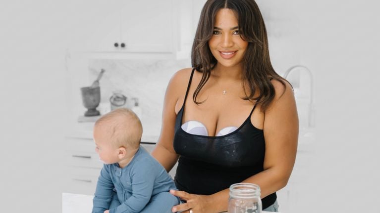 Smiling parent in a black tank top uses two wearable breast pumps while standing beside a baby in a bright kitchen.
