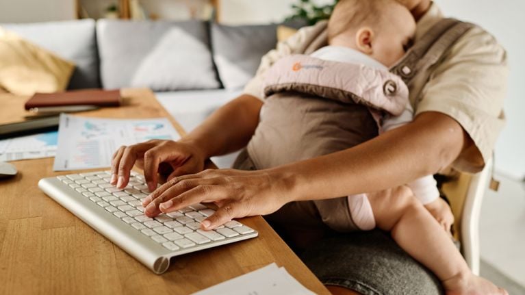 A working parent types on a keyboard at a desk while holding a sleeping baby in a carrier, with papers and a laptop nearby.