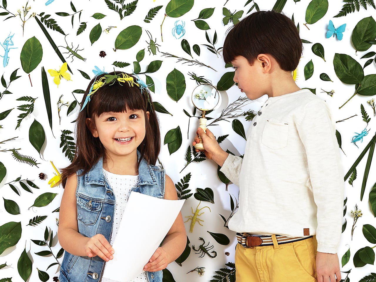 Two kids standing in front of a white wall covered in leaves and toy bugs