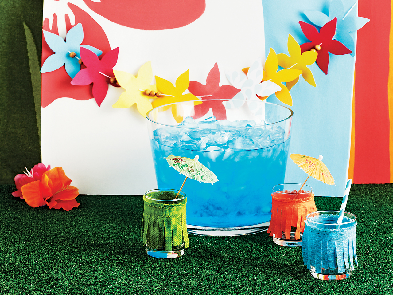 Glasses of blue punch with little umbrellas in front of a garland made to look like a Hawaiian lei