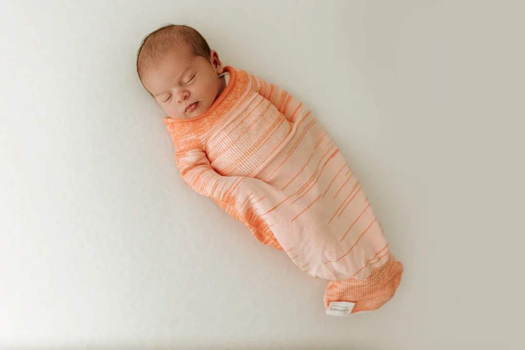A baby in an orange swaddle lies on flat mattress