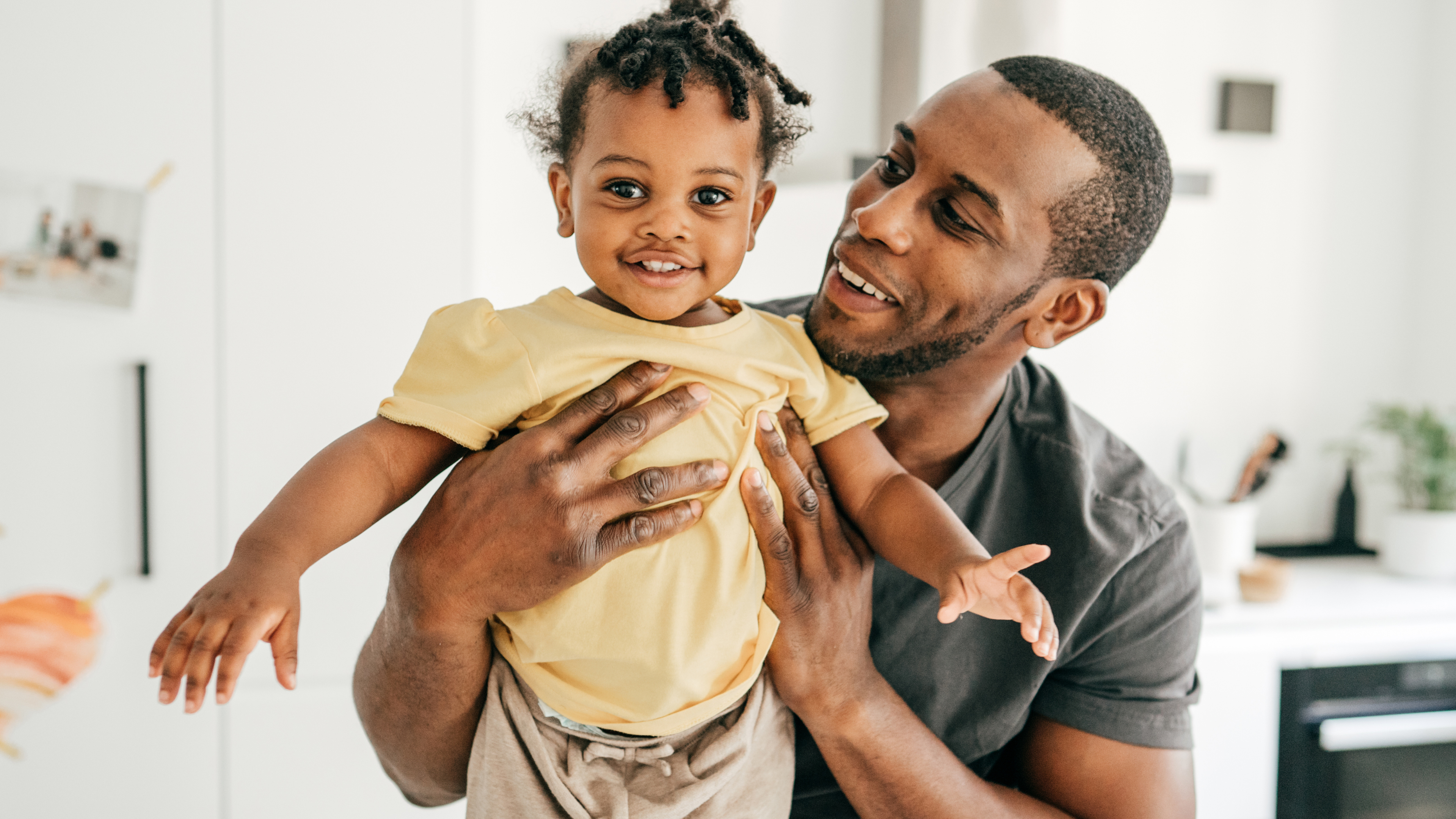 A smiling father lifts his cheerful toddler daughter in the air. The child is wearing a yellow shirt and looking directly at the camera, while the father looks at her with joy in a bright, modern home setting.