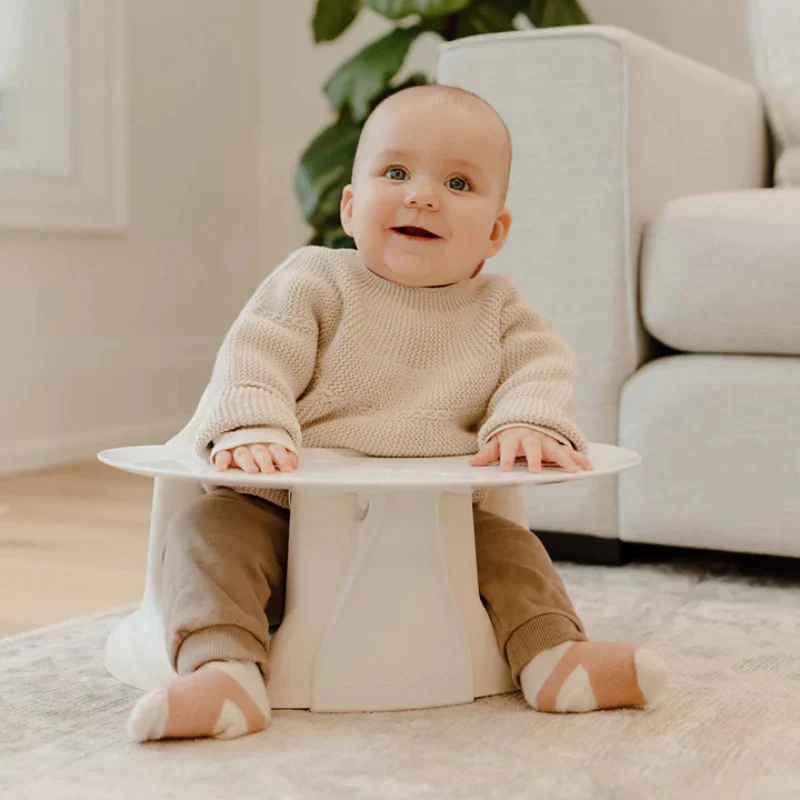 A baby in a beige sweater and brown pants sits in a floor seat with an attached tray, smiling in a cozy living room with soft lighting and neutral decor.