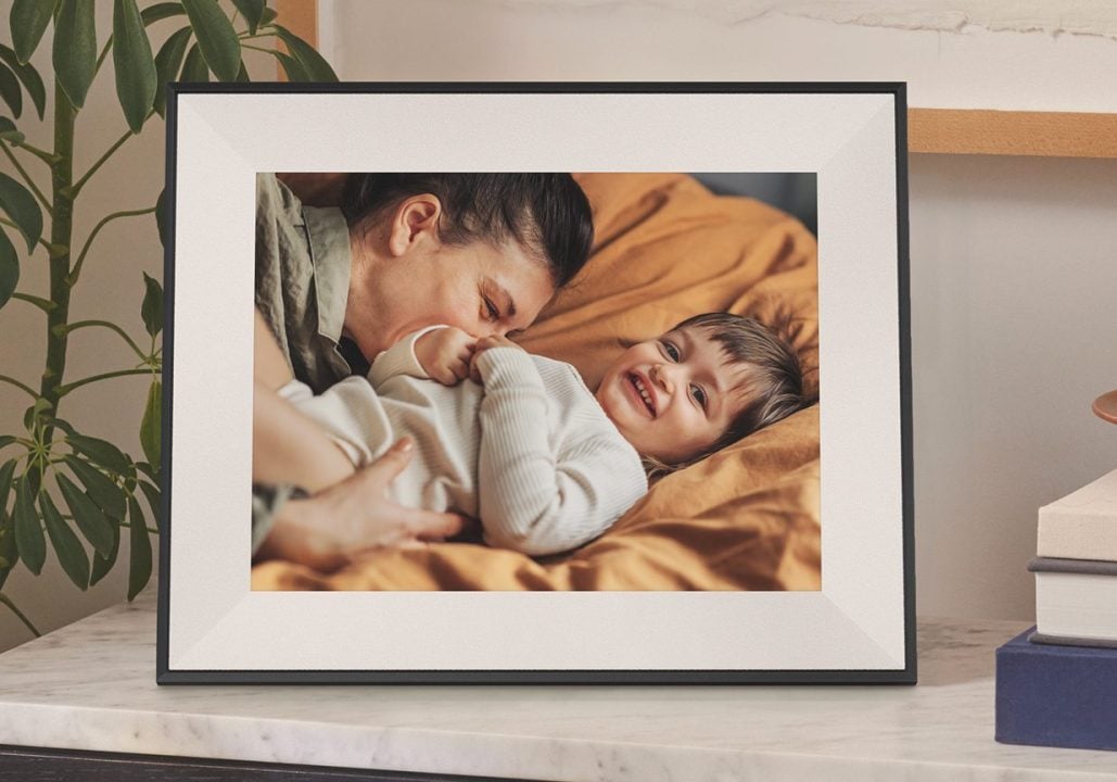 The new Aspen digital frame from Aura displays a photo of a smiling toddler and an adult on a cozy bed, set atop a marble cabinet with books, a dish, and a leafy plant in the background.
