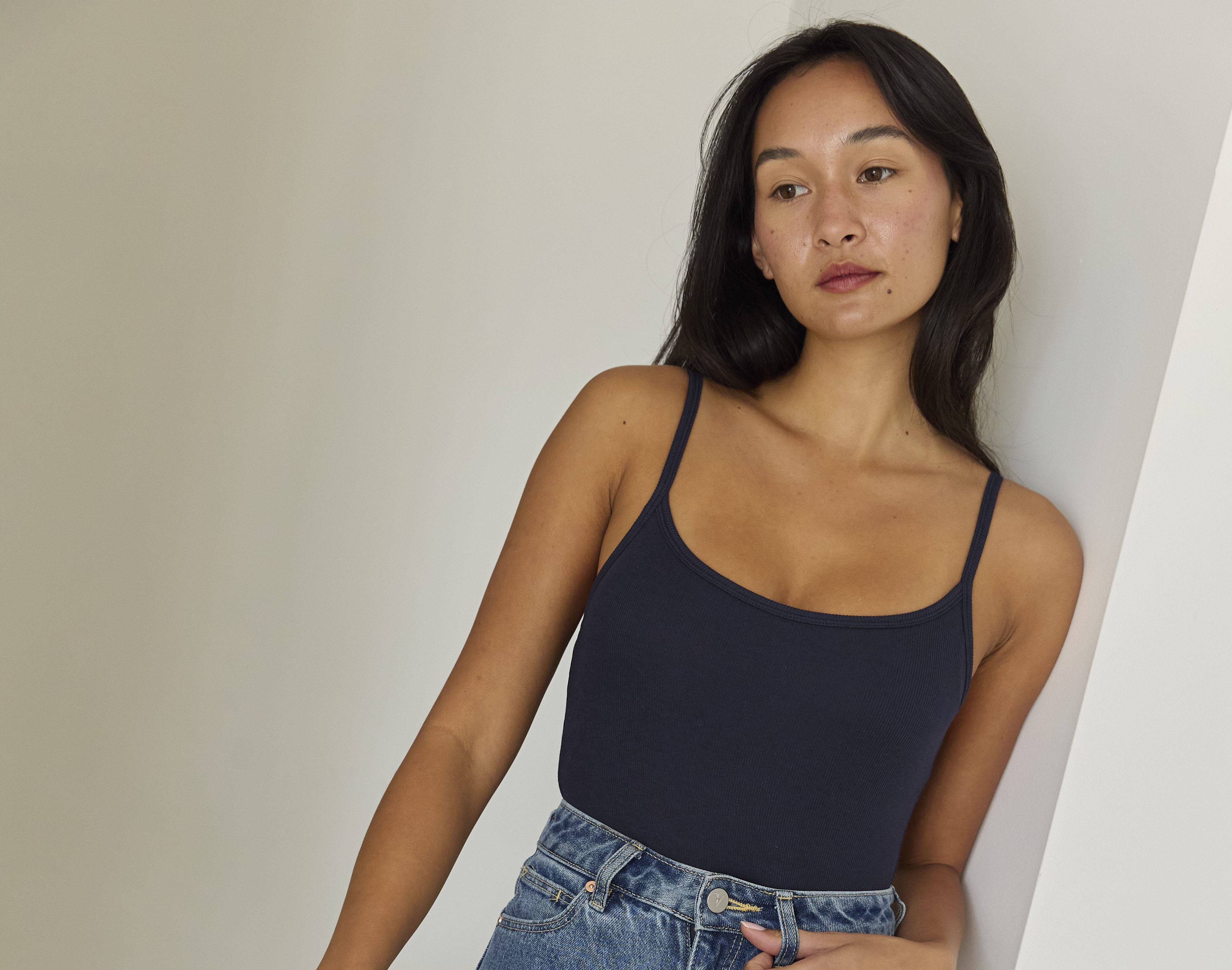 A woman poses indoors in an Embrace navy scoop-neck tank top and high-waisted, relaxed-fit blue jeans, standing barefoot with one knee raised and resting against a white wall.