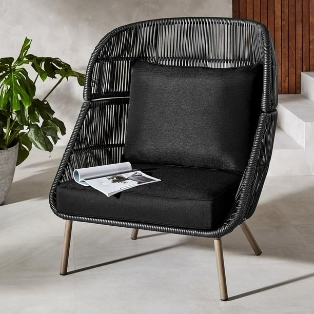 A black woven lounge chair with sleek metal legs and thick cushions sits on a light stone floor, with an open magazine resting on the seat and a leafy green plant nearby.