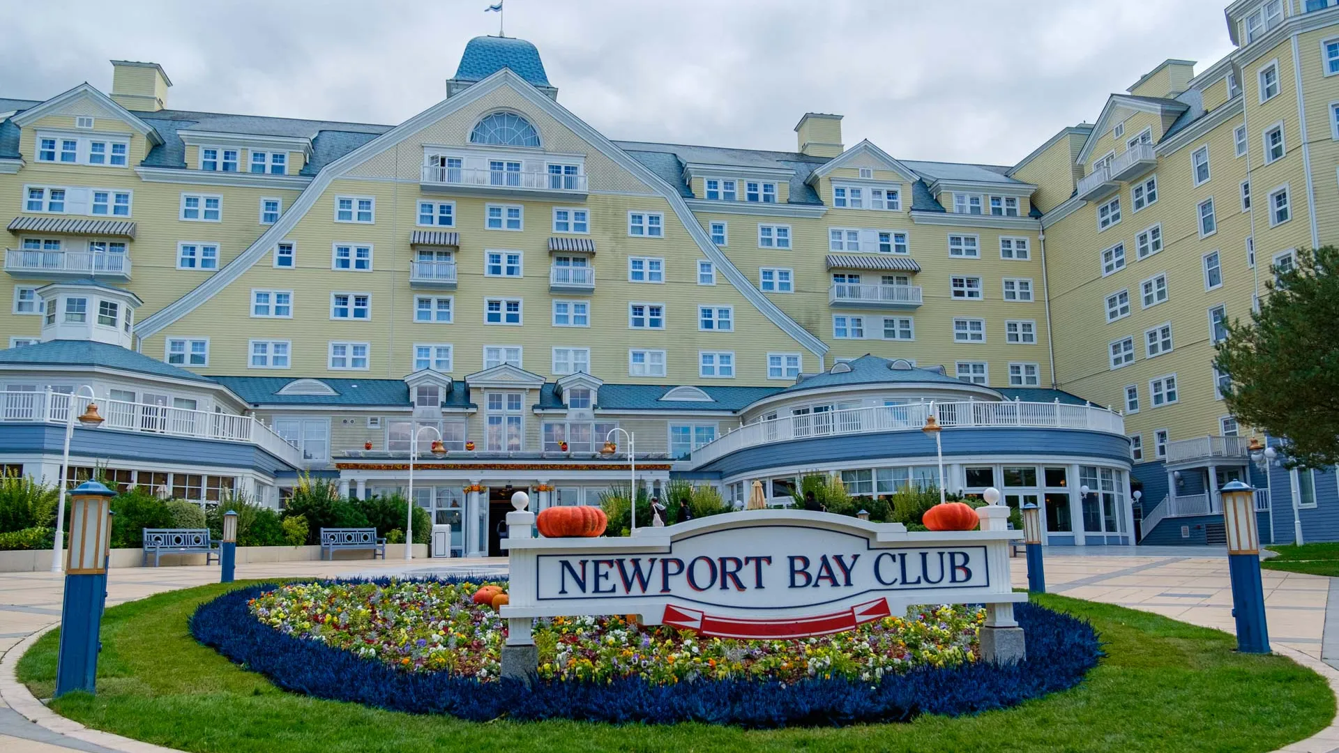 Exterior view of Disney’s Newport Bay Club hotel, a large yellow and blue New England–style building with white trim, multiple windows, and a central arched roof. In front, a landscaped flower bed with pumpkins surrounds a sign reading ‘Newport Bay Club.