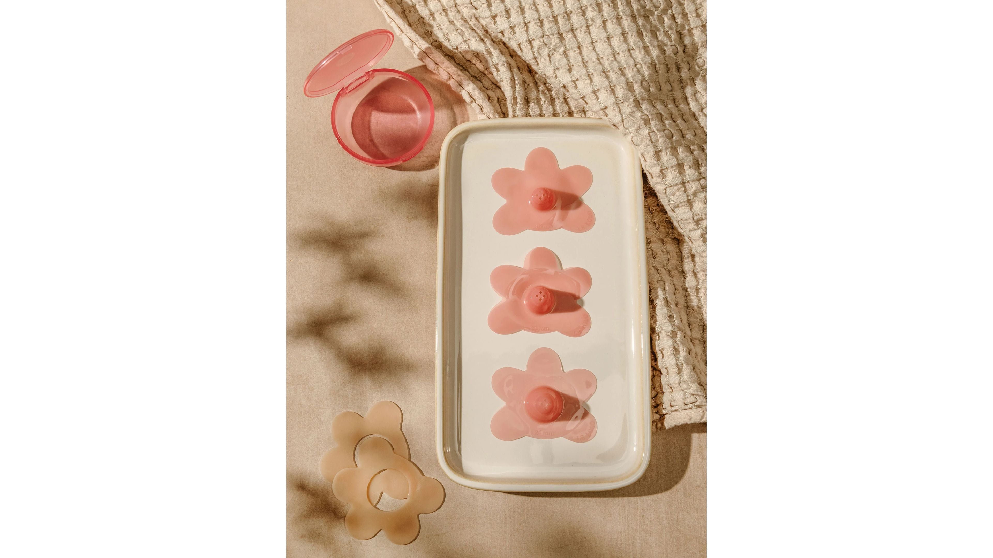 Four pink silicone nipple shields with flower-shaped bases are arranged on a white tray next to a pink storage case and a beige textured cloth.