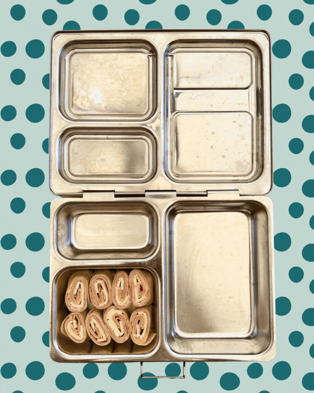 Animated GIF of a metal lunchbox being packed with turkey and cheese pinwheels, fresh berries, a yogurt cup decorated with a smiley face made of fruit, and two snack bars. The background features a teal polka dot pattern on a light green backdrop.