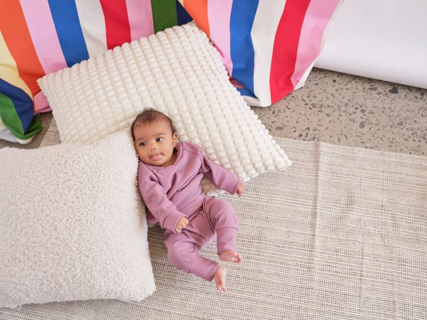 Baby in a mauve one-piece outfit lying on a floor mat, surrounded by textured cream pillows and colorful striped cushions.