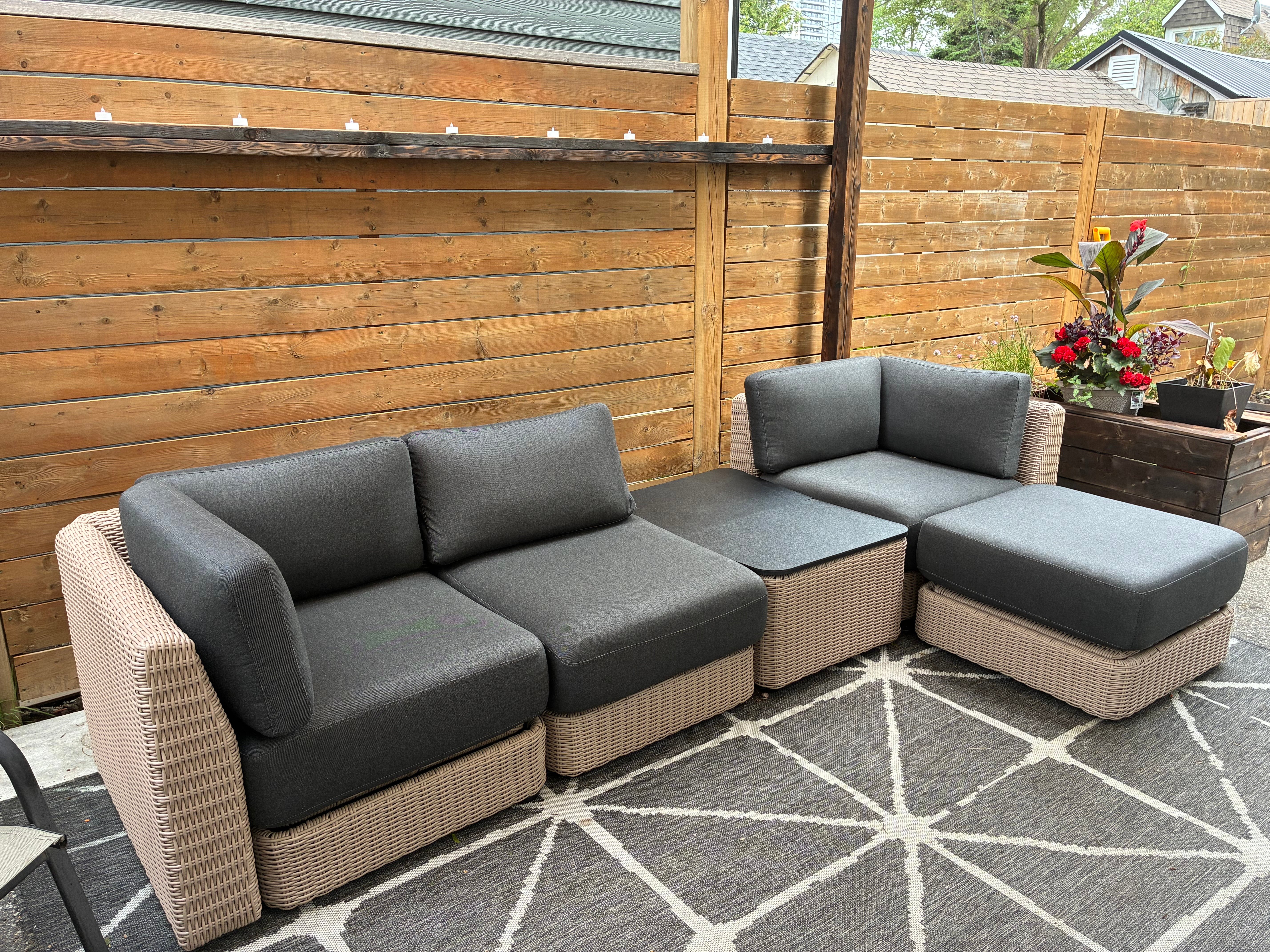 Modular wicker patio sectional with dark charcoal cushions arranged on an outdoor rug in a fenced backyard.